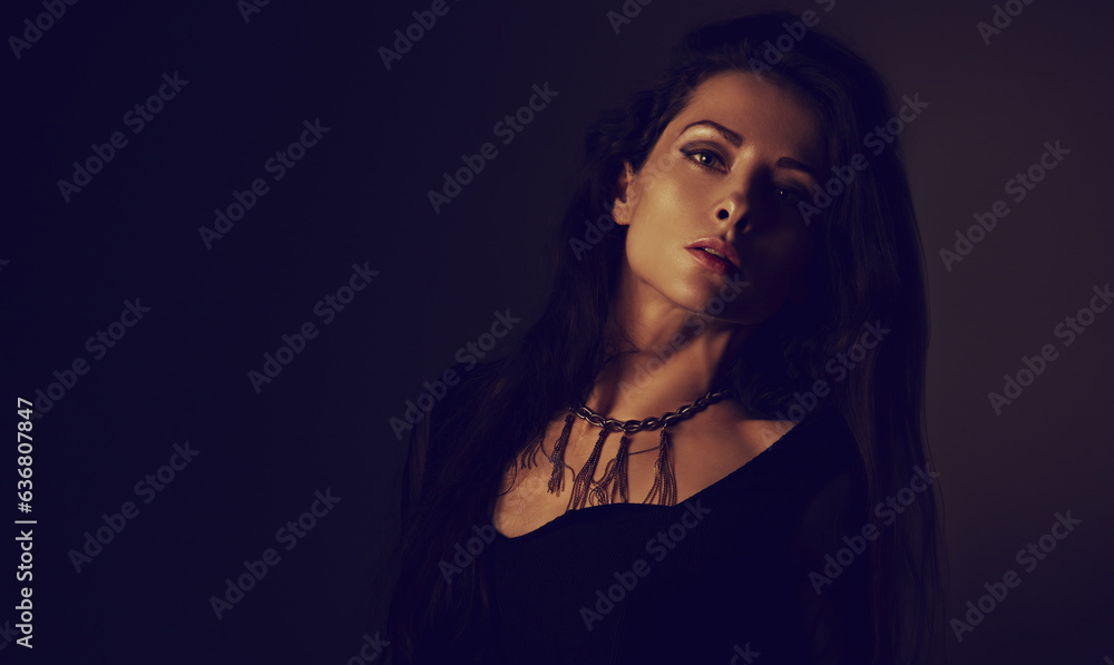 Fototapeta premium Beautiful woman in black dress posing in fashion necklace on the neck. Long hairstyle on dark shadow background. Toned portrait. Closeup.