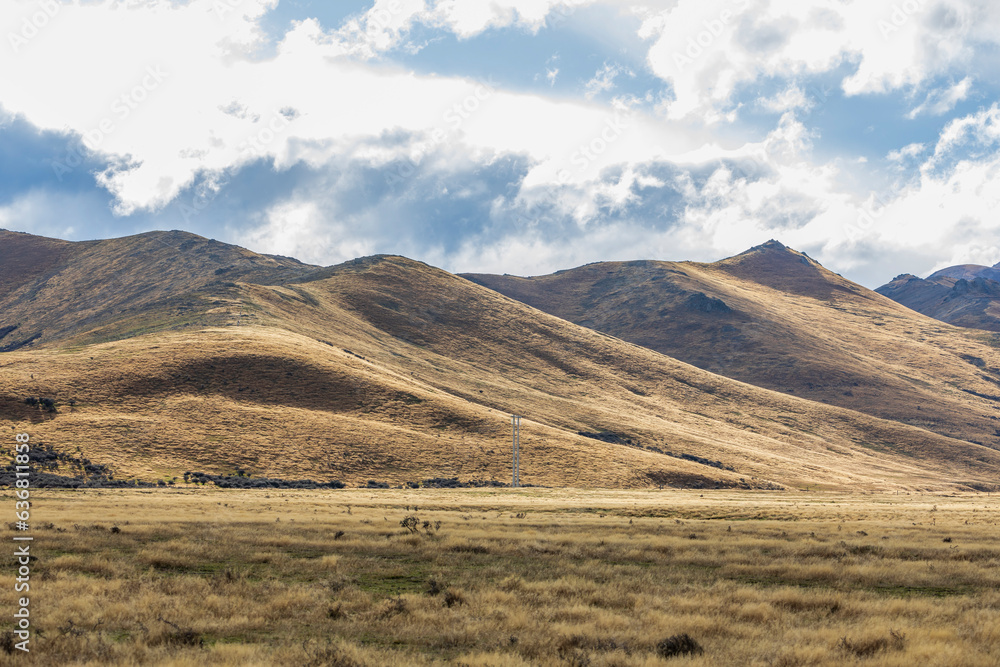 Photograph of a dry mountain range running behind a large brown agricultural field with low level grey clouds on the South Island of New Zealand