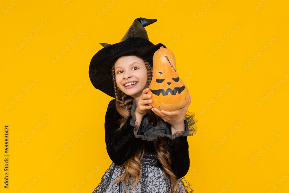 A little witch in a pointed hat holds an evil pumpkin. A child in a festive carnival costume on halloween smiles happily. Children's party. Yellow isolated background.