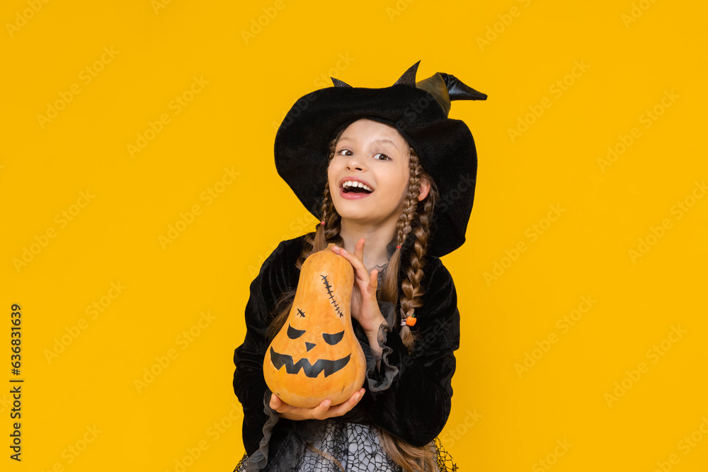 A little witch in a pointed hat holds an evil pumpkin. A child in a festive carnival costume on halloween smiles happily. Children's party. Yellow isolated background. A little witch in a pointed hat holds an evil pumpkin. A child in a festive carnival costume on halloween smiles happily. Children's party. Yellow isolated background.