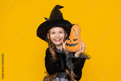 A little girl in a witch costume holds an evil pumpkin in her hands. Children's holiday carnival on Halloween. A happy child rejoices in a pointed black wizard hat. Yellow isolated background.