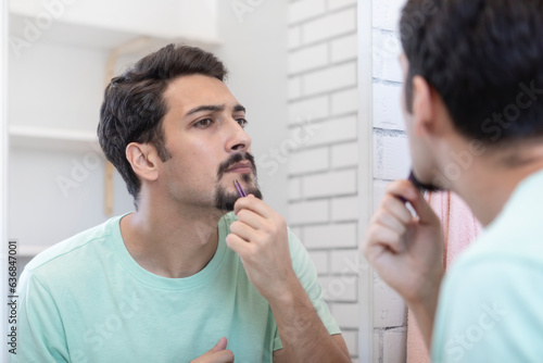 Handsome man grooming in front of the mirror in the bathroom. Young guy depilates with tweezers