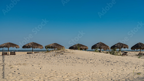A row of straw sun umbrellas and sunbeds on the beach. Clear blue sky. Turquoise ocean ahead. Footprints in the sand. Madagascar. Morondava. © Вера 
