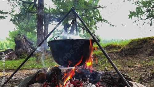 Tourists prepare lunch in a camping pot on a campfire. Cooking in the forest on the banks of a mountain river outdoors. Travel camp, campfire site. Slow motion video.