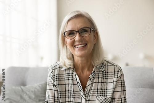 Happy positive blonde senior woman in eye glasses looking at camera with toothy smile, laughing, speaking, sitting on sofa. Older freelance business lady head shot video call portrait