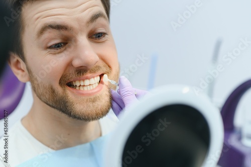 Dentist in blue medical gloves applying sample from tooth enamel scale to smiling man patient teeth to pick up right shade, teeth bleaching procedure