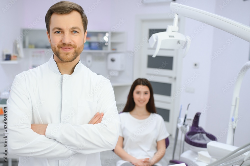 Portrait of positive male dentist with crossed hands posing at camera with patient and nurse on background Portrait of positive male dentist with crossed hands posing at camera with patient and nurse on background