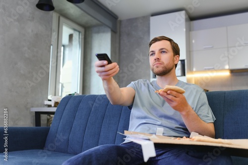 Caucasian man resting on sofa, eating pizza and watching television. Concept of modern successful man's lifestyle and relax after work. Comfortable studio apartment.