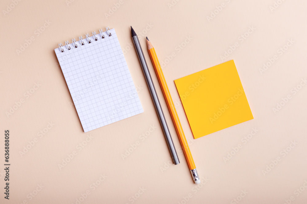 Stationery from above. Checkered notepad mockup, orange and grey pencils, orange paper for notes on light beige background top view. Place for text. Stationery from above. Checkered notepad mockup, orange and grey pencils, orange paper for notes on light beige background top view. Place for text.