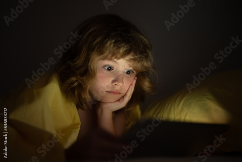 Cute little kid lying under blanket with tablet. Surprised kid in bed under blanket watching video on tablet in dark bedroom with glowing lights. Kid on bed at night watching movie.