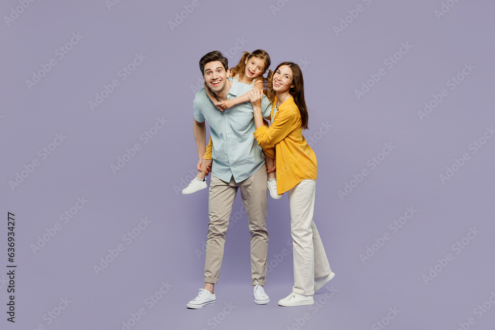 Full body young joyful happy parents mom dad with child kid daughter girl 6 years old wear blue yellow casual clothes giving piggyback ride to joyful, sit on back isolated on plain purple background. Full body young joyful happy parents mom dad with child kid daughter girl 6 years old wear blue yellow casual clothes giving piggyback ride to joyful, sit on back isolated on plain purple background.