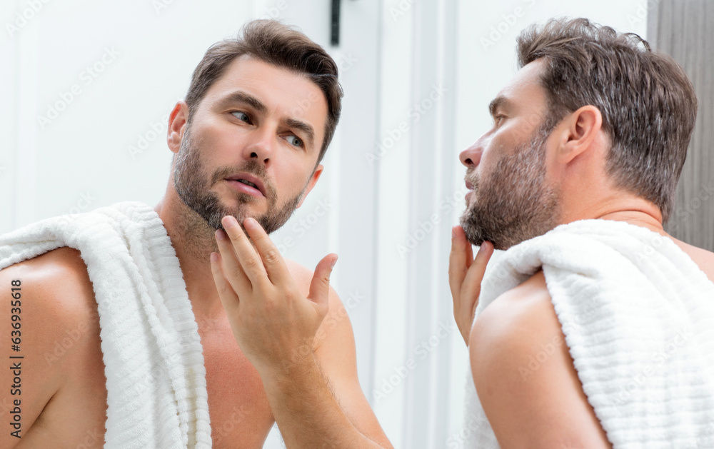 Fototapeta premium Man applying cosmetic face cream on mirror. Facial treatment. Portrait of man with bare naked shoulders touching skin. Sexy man with skin care product. Male face creme. Perfect skin, morning routine.