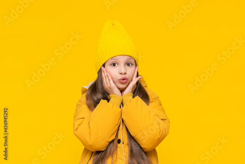A little girl in a beanie hat and an orange jacket pursed her lips in surprise. Warm autumn clothes for teenagers and children. The child is surprised. Yellow isolated background.