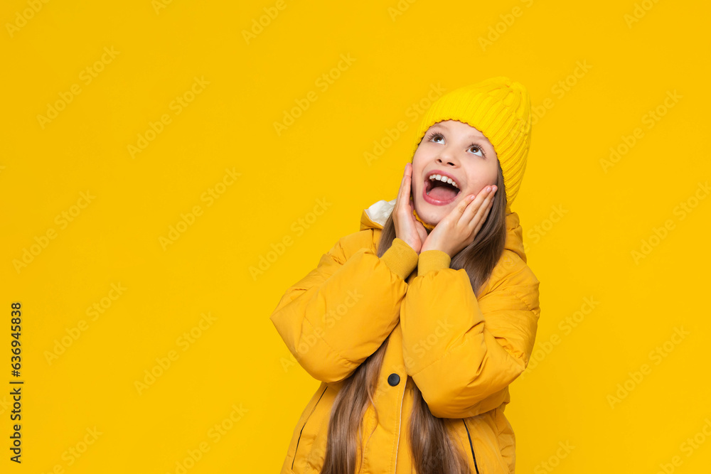 A happy little girl in a jacket and hat. Autumn children's fashion. A young girl in a warm down jacket grabbed her cheeks and opened her mouth wide. Yellow isolated background. Copy space. Banner. A happy little girl in a jacket and hat. Autumn children's fashion. A young girl in a warm down jacket grabbed her cheeks and opened her mouth wide. Yellow isolated background. Copy space. Banner.