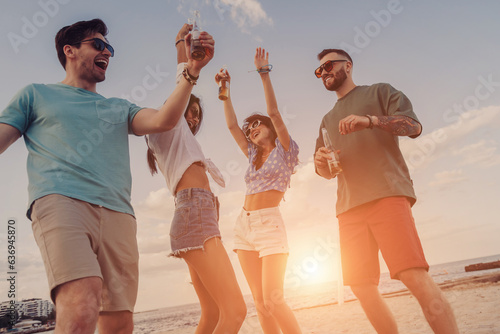 Four cheerful young people enjoying cold beer while dancing on the beach together