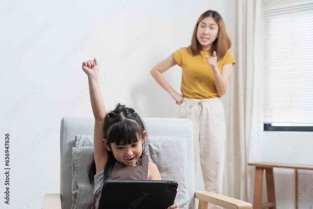 Asian mother looking at her daughter playing game on tablet while studying Asian mother looking at her daughter playing game on tablet while studying