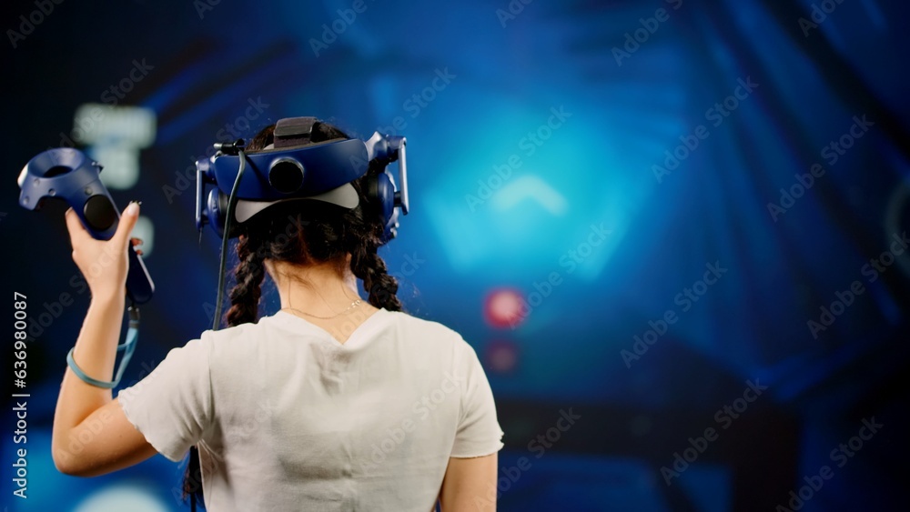 The child use tracking controllers in her hands, moves actively immersing herself in modern game. The little girl wearing VR headset plays game. Behind girl there is screen with blur background image The child use tracking controllers in her hands, moves actively immersing herself in modern game. The little girl wearing VR headset plays game. Behind girl there is screen with blur background image