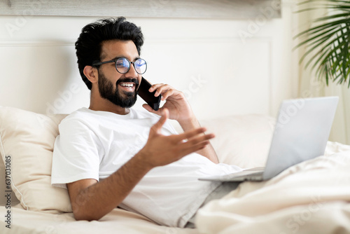 Indian man talking on cellphone and using laptop while relaxing in bed