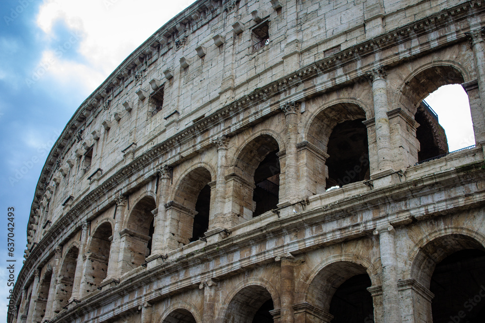 A close-up view of the Colosseum's exterior, highlighting the intricate details of the arches and stonework, with a hint of the Roman sky peering through, capturing the essence of ancient grandeur.
