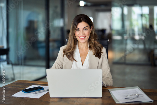 Happy smiling mature mid aged professional business woman manager executive or lawyer attorney looking at camera at workplace working on laptop computer technology in office sitting at desk, portrait. photo