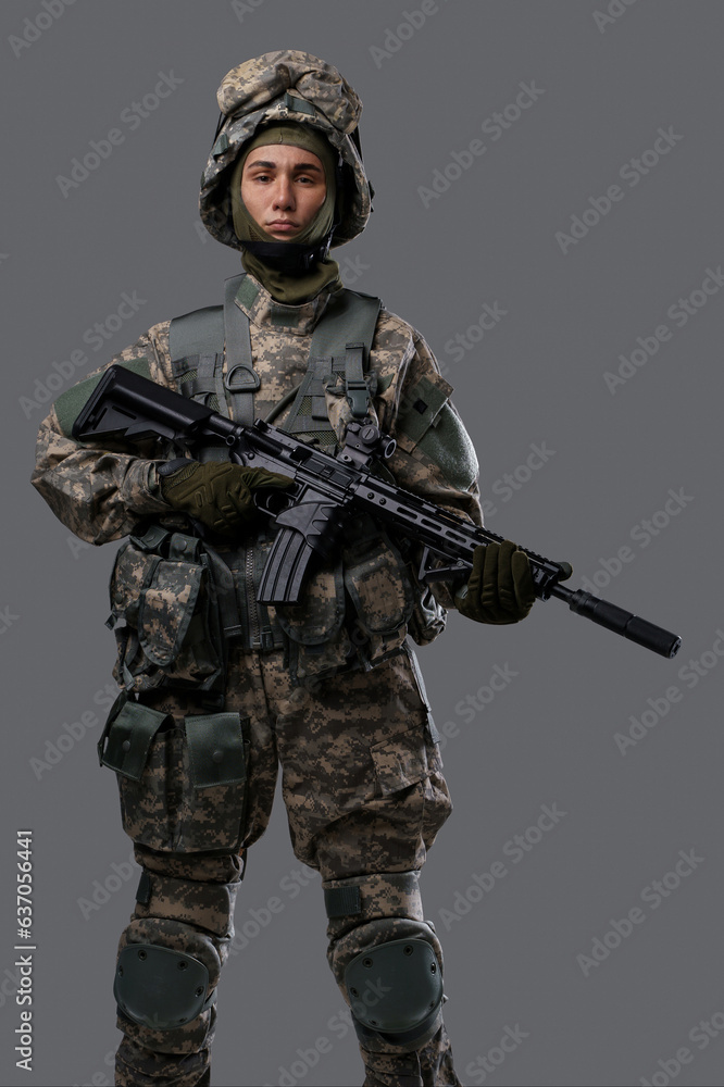 Young man in military uniform and helmet holding a rifle on a grey background Young man in military uniform and helmet holding a rifle on a grey background