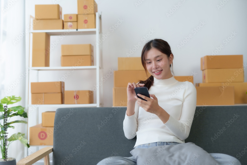 Asian businesswoman starting sme sits on the sofa using mobile phone at home. Small Online Business Owner, SME Work From Home Box, SME Online Marketing and Product Packing and Delivery Service. Asian businesswoman starting sme sits on the sofa using mobile phone at home. Small Online Business Owner, SME Work From Home Box, SME Online Marketing and Product Packing and Delivery Service.
