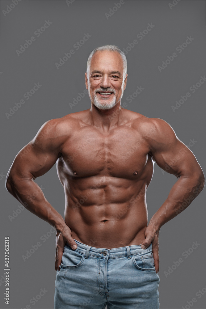 Attractive older man with a muscular physique, stylish gray beard and ripped jeans, smiling and posing shirtless against a gray backdrop Attractive older man with a muscular physique, stylish gray beard and ripped jeans, smiling and posing shirtless against a gray backdrop