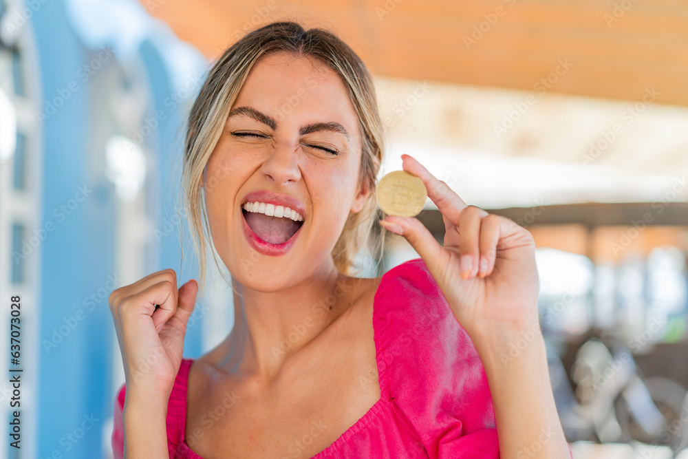 Young blonde woman at outdoors holding a Bitcoin and celebrating a victory Young blonde woman at outdoors holding a Bitcoin and celebrating a victory