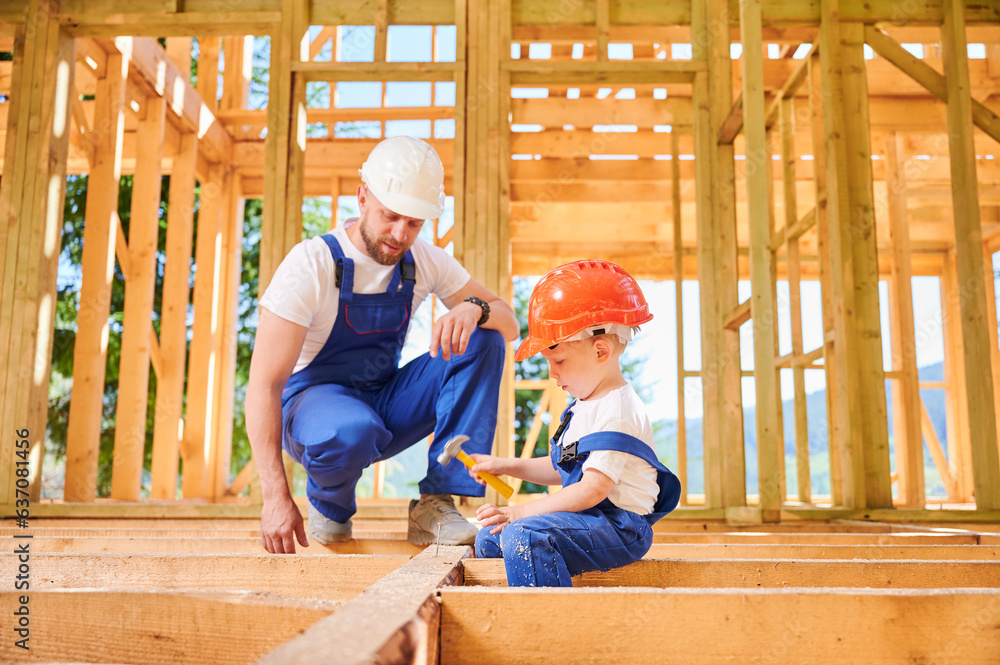 Father with toddler son building wooden frame house. Worker instructing