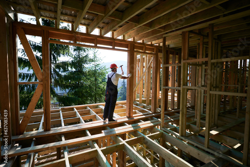 Carpenter constructing wooden frame, two-story house near forest. Bearded man hammering nails with hammer, dressed in protective helmet and work coveralls. Concept of modern ecological construction. © anatoliy_gleb