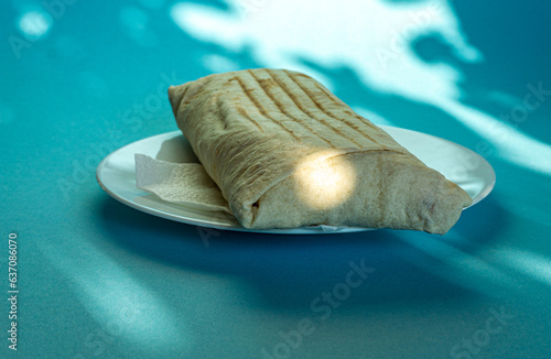Large shawarma on a white plate with a napkin on a blue background with shadows. Food, healthy fast food, delicious to eat.