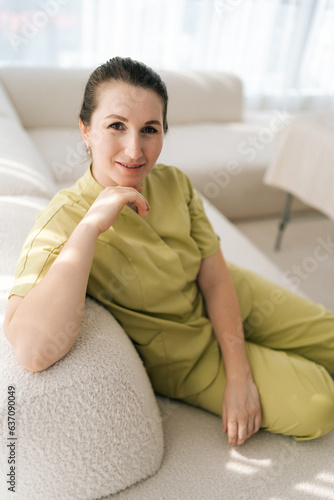 Closeup vertical portrait of female masseuse therapist in uniform sitting posing on couch looking at camera with friendly expression, on background of window in wellness center. Concept of body care.