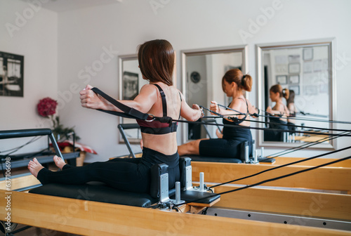 Two fit-shaped females doing back strength exercises using pilates reformer machine in the sport athletic gym with a mirror wall. Active people training or medical rehabilitation concept image. © Soloviova Liudmyla
