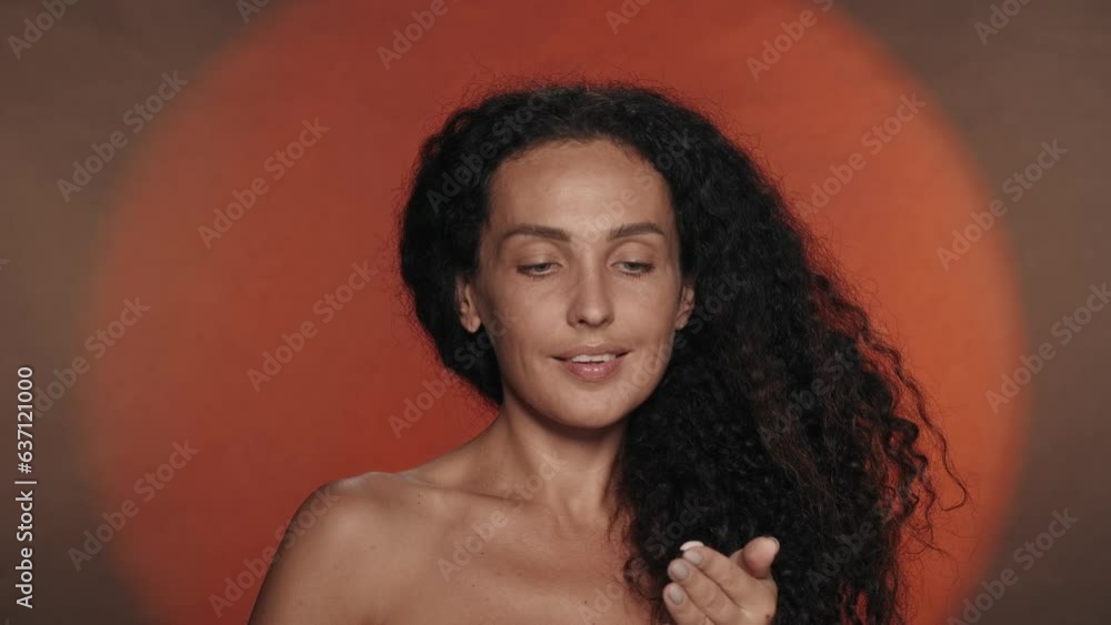 Hand care and protection. A woman applies cream from a black bottle on her hands, moisturizing her skin. Portrait of a seminude curly woman in the studio on an orange background with circular light. Hand care and protection. A woman applies cream from a black bottle on her hands, moisturizing her skin. Portrait of a seminude curly woman in the studio on an orange background with circular light.