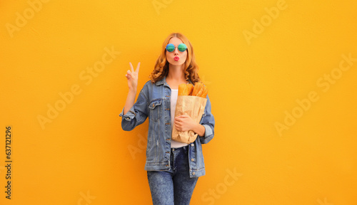 Portrait of stylish happy young woman posing with grocery shopping paper bag with long white bread baguette on orange background