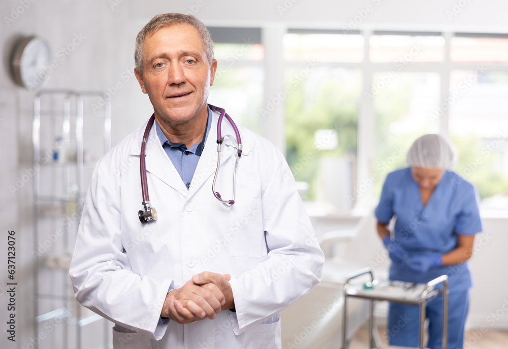 Fototapeta premium Experienced smiling elderly male doctor in white coat standing in modern light medical office with female assistant in blue uniform preparing instruments in background