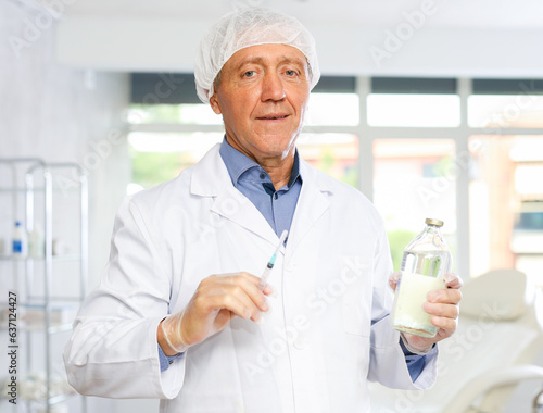 Experienced aged male doctor in white coat, medical cap and gloves standing in clinic office with syringe in hands, ready to make injection or administer medication or vaccine to patient