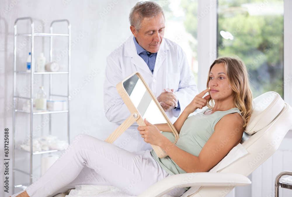 Obraz premium Female patient of cosmetology office sitting in chair with mirror in hands and discussing with mature male doctor need for rejuvenation procedure