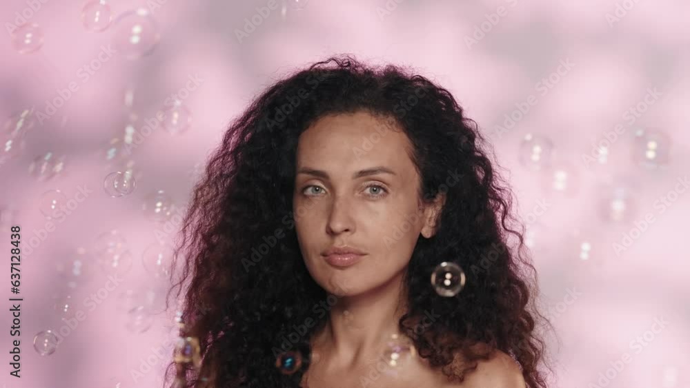 Portrait of a seminude woman in the studio on a pink background with highlights close up. The woman brushes off the soap bubbles swirling around her. The concept of beauty, cosmetology, skin care. Portrait of a seminude woman in the studio on a pink background with highlights close up. The woman brushes off the soap bubbles swirling around her. The concept of beauty, cosmetology, skin care.