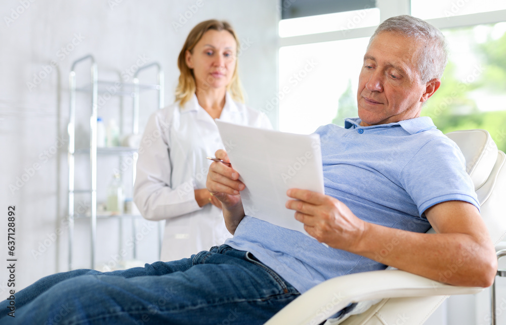 Obraz premium Before medical manipulation, mature man patient sits in chair and reads documents with woman doctor about upcoming manipulation. Medical documentation, questionnaire, patient survey