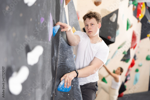 Teenage boy is mastering climbing on training wall in gym, side view. Teen guy holds on tightly to ledges and strives for top of bouldering route
