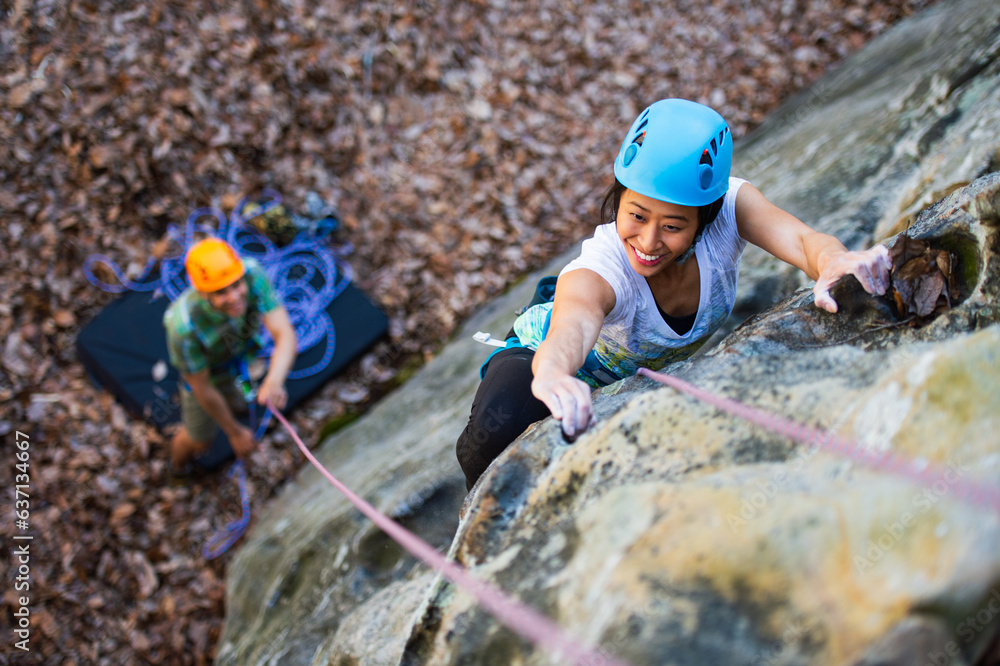 A young asian woman in a blue helmet climbs a rock cliff on top rope while her partner wearing an orange helmet belays from below, seen from above A young asian woman in a blue helmet climbs a rock cliff on top rope while her partner wearing an orange helmet belays from below, seen from above