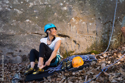 A young woman sitting in front of a sandstone cliff smiles while putting on climbing shoes before a rock climb photo