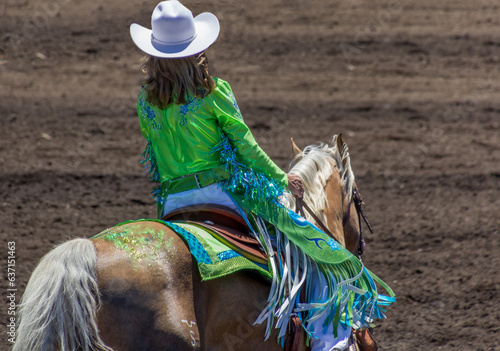 A cowgirl at a rodeo is wearing in green costume and sitting on the back of a blond horse. She has on a white hat. The horse has green glitter on its back. The arena is dirt.