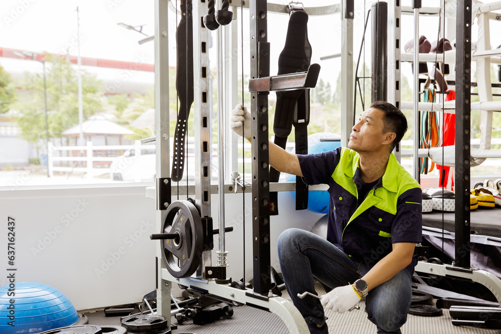 Professional Asian male service worker or fitter checks equipment, maintains and secures fitness equipment in indoor gym, provides safety for users : Skilled technicians repair exercise machines. - obrazy, fototapety, plakaty 