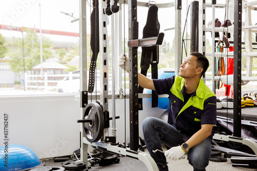 Professional Asian male service worker or fitter checks equipment, maintains and secures fitness equipment in indoor gym, provides safety for users : Skilled technicians repair exercise machines. photo