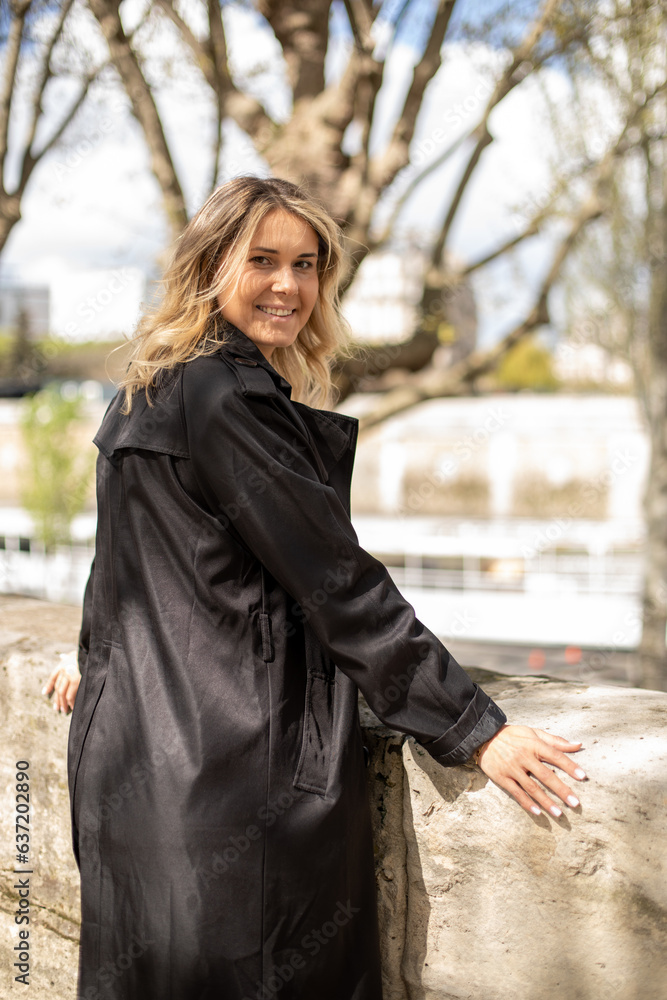 Side view of young smiling woman with fair hair wearing black trench coat, putting hands on concrete fence near river. Side view of young smiling woman with fair hair wearing black trench coat, putting hands on concrete fence near river.