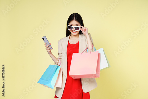 Young woman holding shopping bags while using mobile phone isolated over yellow background.