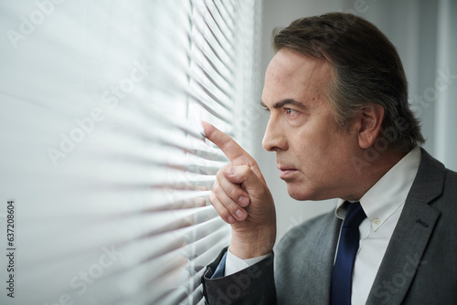 Curious mature businessman peaking through window blinds