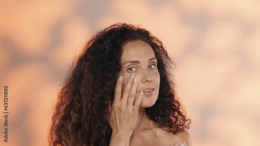 A woman touches her face, enjoying smooth skin after a cosmetic procedure. Portrait of a semi-nude woman in the studio on an orange background with highlights.